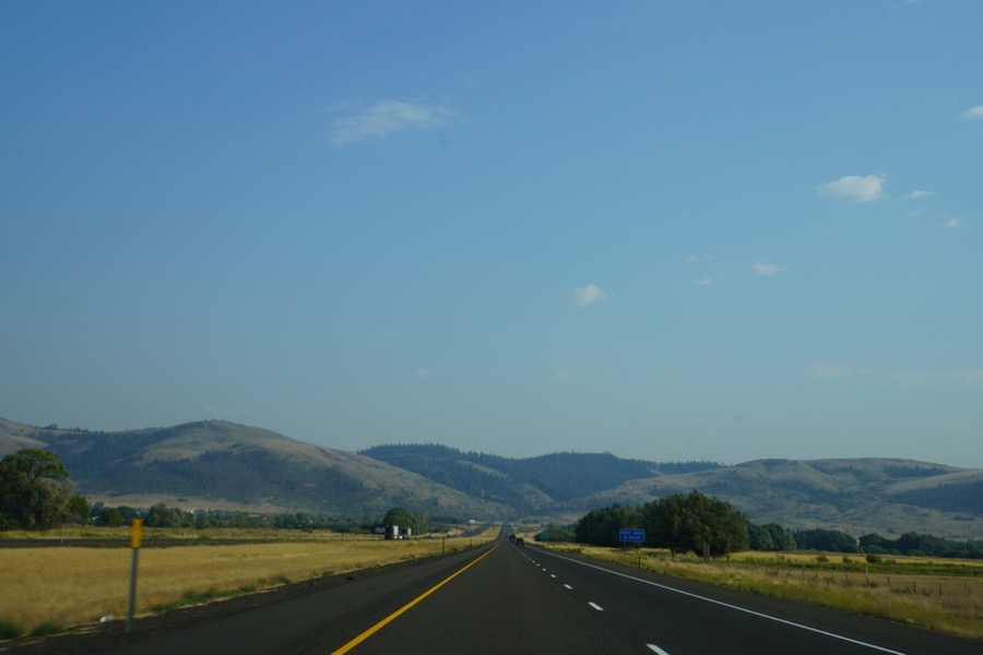 A two-lane highway crossing open fields toward low, dry hills.