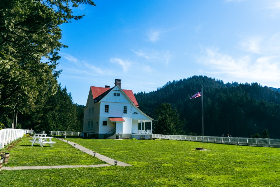 A white house with a red roof sits in a lawn below a wooded hillside.